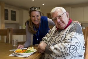 Community Care Worker Serves Up Lunch To A Man Sitting At The Kitchen Table